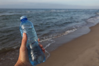 Photo of Woman holding plastic bottle with water near sea, closeup. Space for text