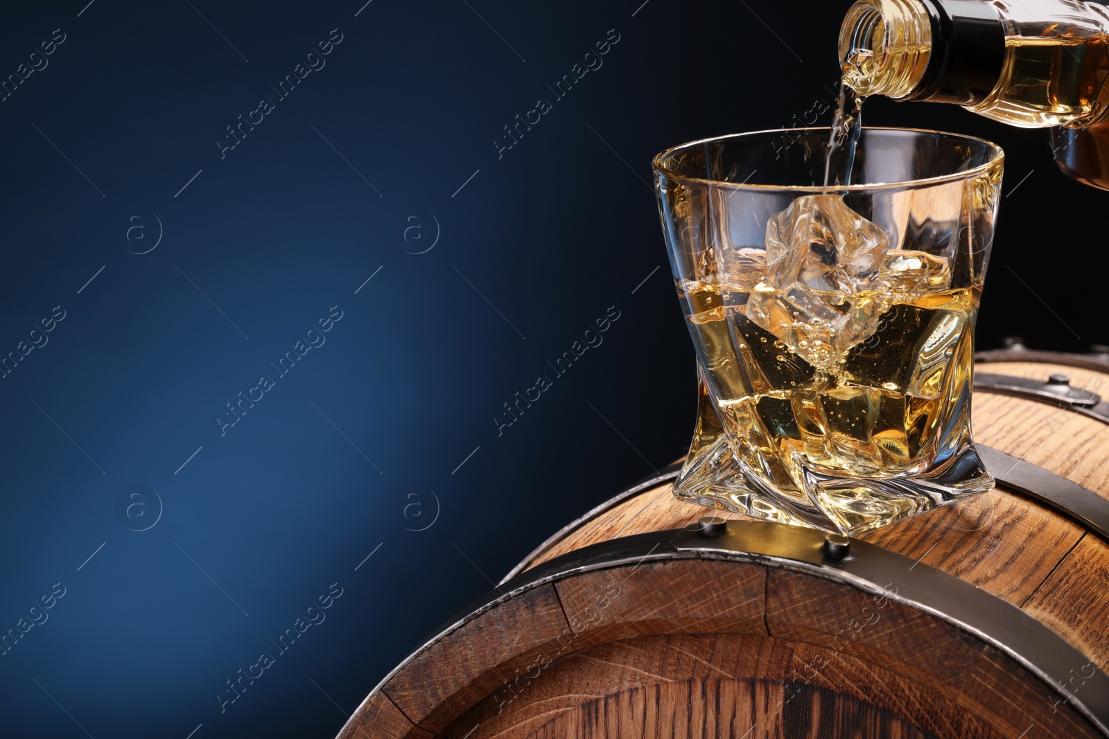 Photo of Pouring whiskey from bottle into glass on wooden barrel against blue background, closeup. Space for text