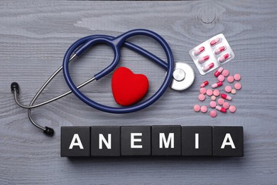 Photo of Word Anemia made with black cubes, stethoscope, pills and decorative heart on grey wooden table, flat lay