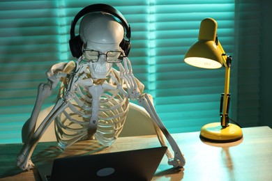 Photo of Waiting concept. Human skeleton sitting at wooden table with laptop and headphones indoors