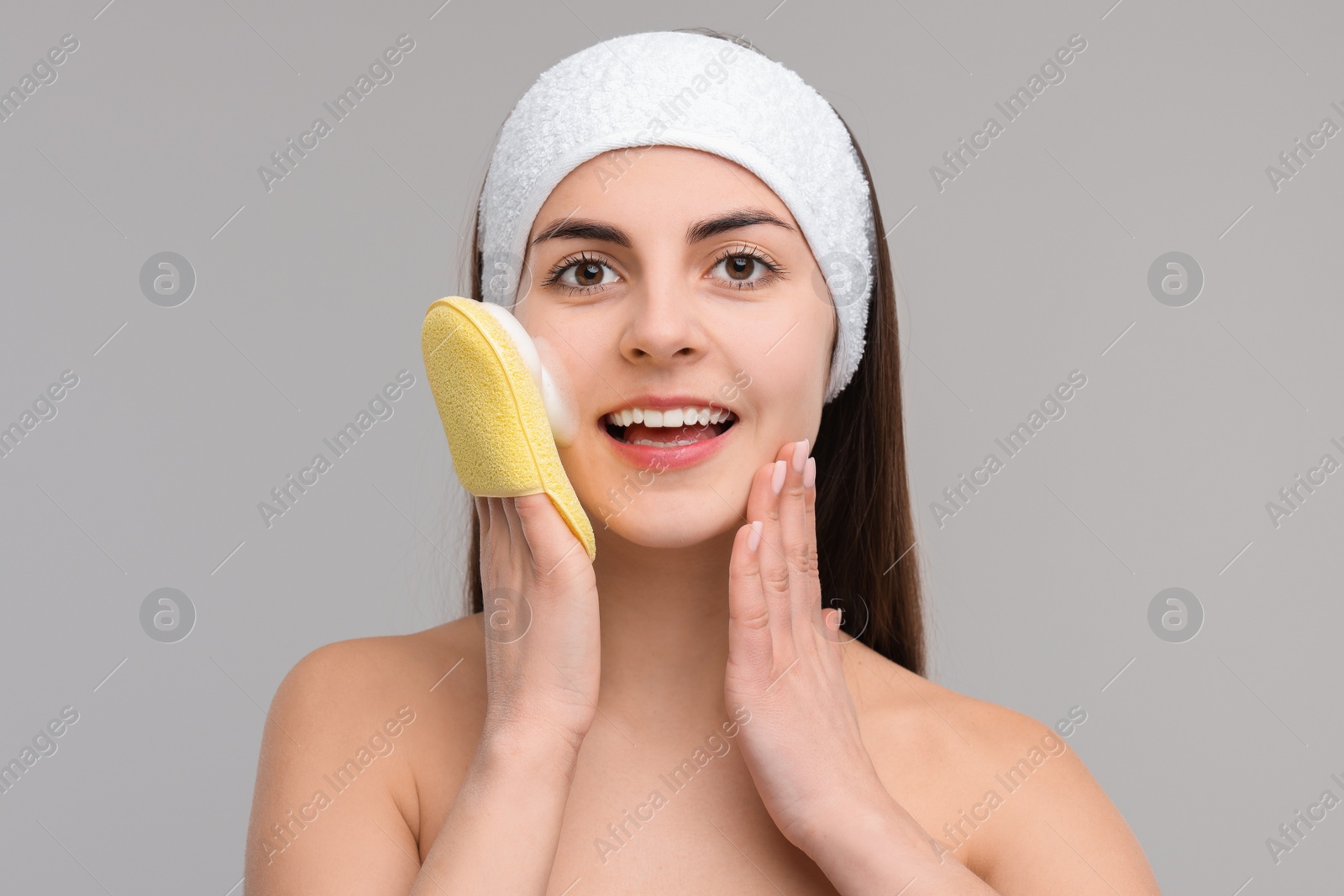 Photo of Young woman with headband washing her face using sponge on light grey background