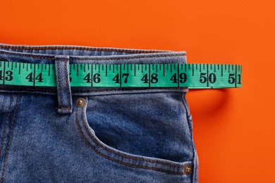 Jeans and measuring tape on orange background, top view. Weight loss concept