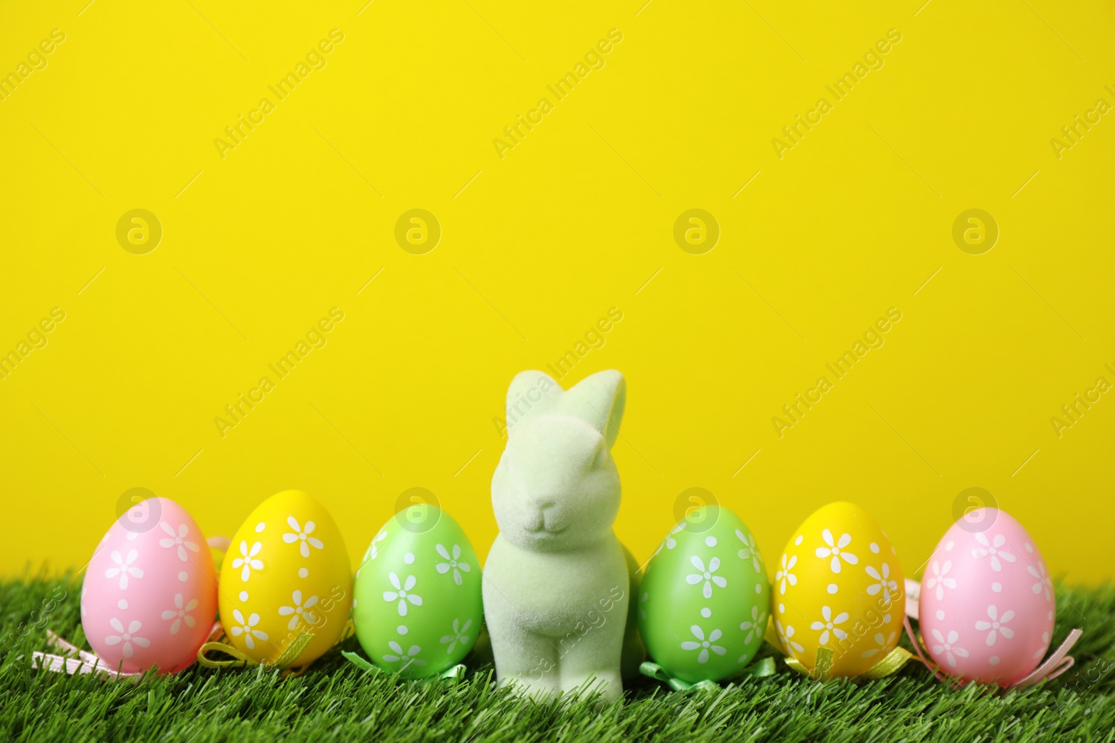 Photo of Easter bunny toy and dyed eggs on green grass against yellow background. Space for text