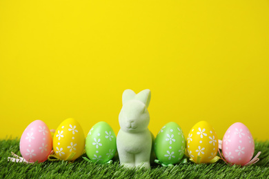 Easter bunny toy and dyed eggs on green grass against yellow background. Space for text