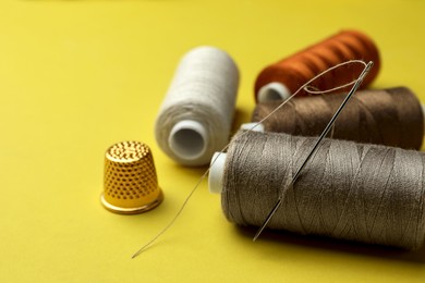 Photo of Different colorful sewing threads with needle and thimble on yellow background, closeup