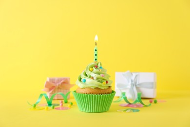 Photo of Delicious birthday cupcake with burning candle, sprinkles and gift boxes on yellow background