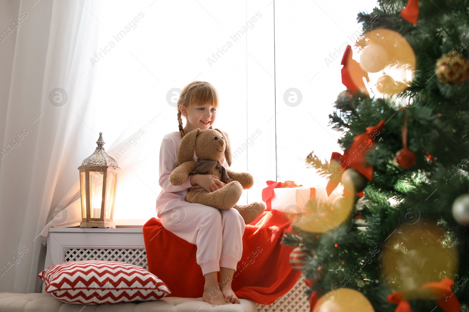 Photo of Cute little child with toy rabbit sitting on windowsill near Christmas tree at home