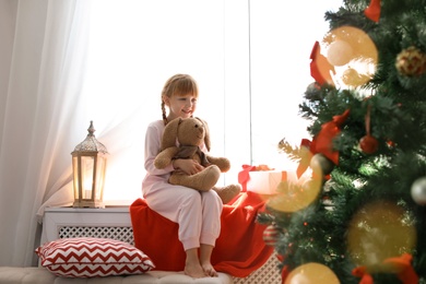 Photo of Cute little child with toy rabbit sitting on windowsill near Christmas tree at home