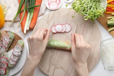 Woman wrapping spring roll at white wooden table with products, top view