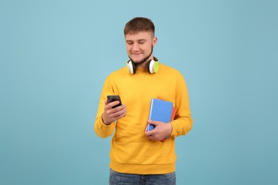 Young student with headphones and books using smartphone on light blue background