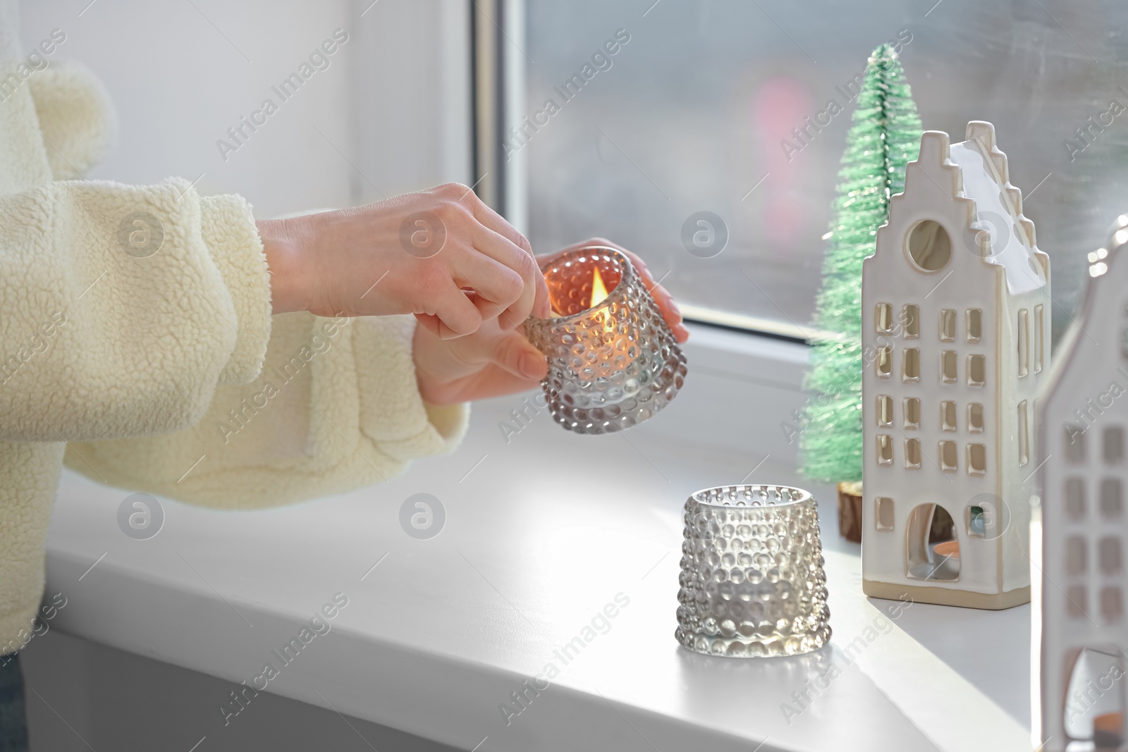 Photo of Woman lightning up candle near windowsill with house shaped lanterns indoors, closeup