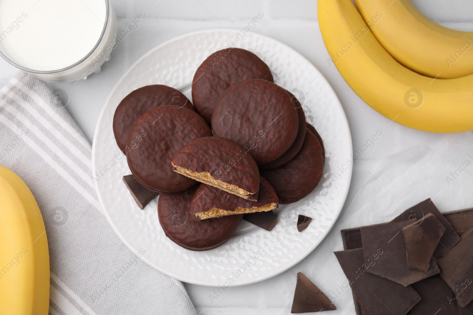 Photo of Tasty banana choco pies, pieces of chocolate, fruits and milk on white tiled table, flat lay