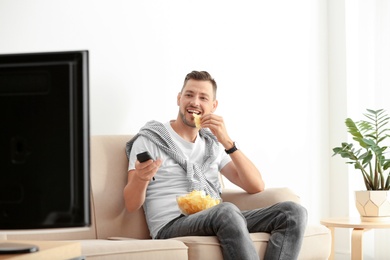 Photo of Man with bowl of potato chips watching TV on sofa in living room