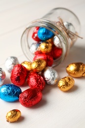 Overturned glass jar with chocolate eggs wrapped in colorful foil on white wooden table