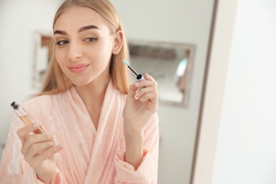 Young woman applying oil onto her eyelashes near mirror indoors