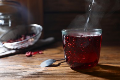 Photo of Making hibiscus tea. Pouring hot water into cup with roselle petals at wooden table, closeup. Space for text