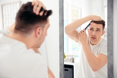 Photo of Young man with hair loss problem looking in mirror indoors