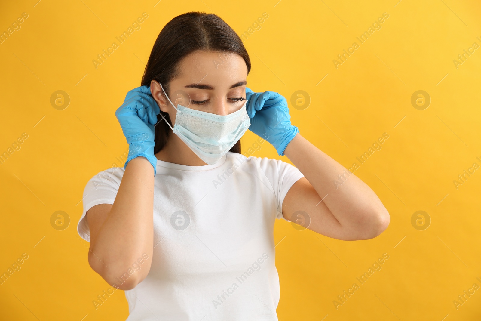 Photo of Woman in medical gloves putting on protective face mask against yellow background