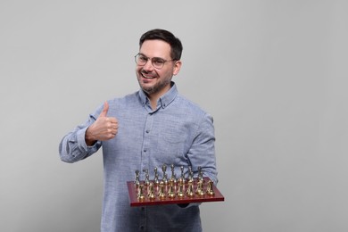Smiling man holding chessboard with game pieces and showing thumbs up on light grey background, space for text