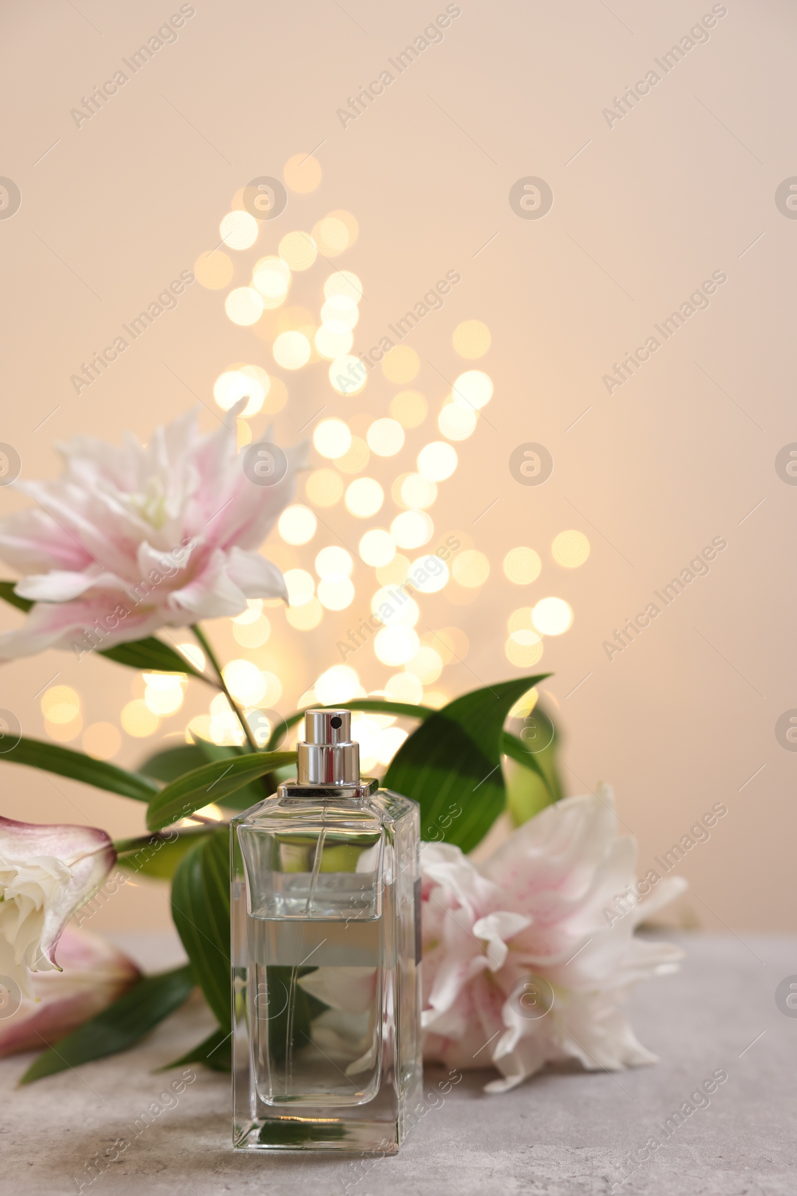 Photo of Bottle of perfume and beautiful lily flowers on table against beige background with blurred lights, space for text