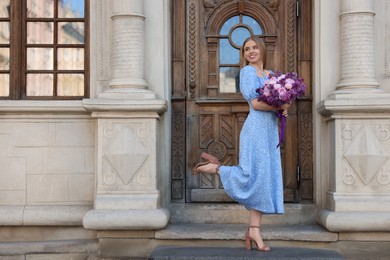 Beautiful woman with bouquet of spring flowers near building outdoors, space for text