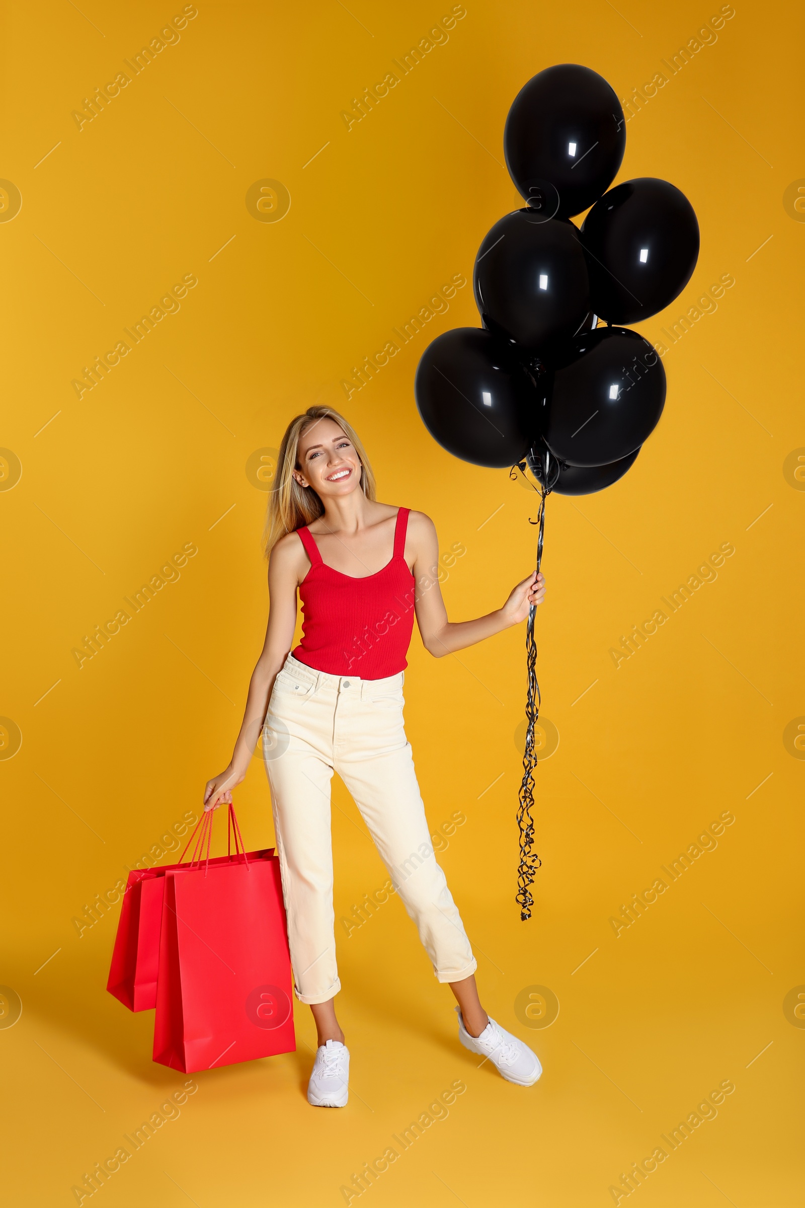 Photo of Happy young woman with balloons and shopping bags on yellow background. Black Friday Sale