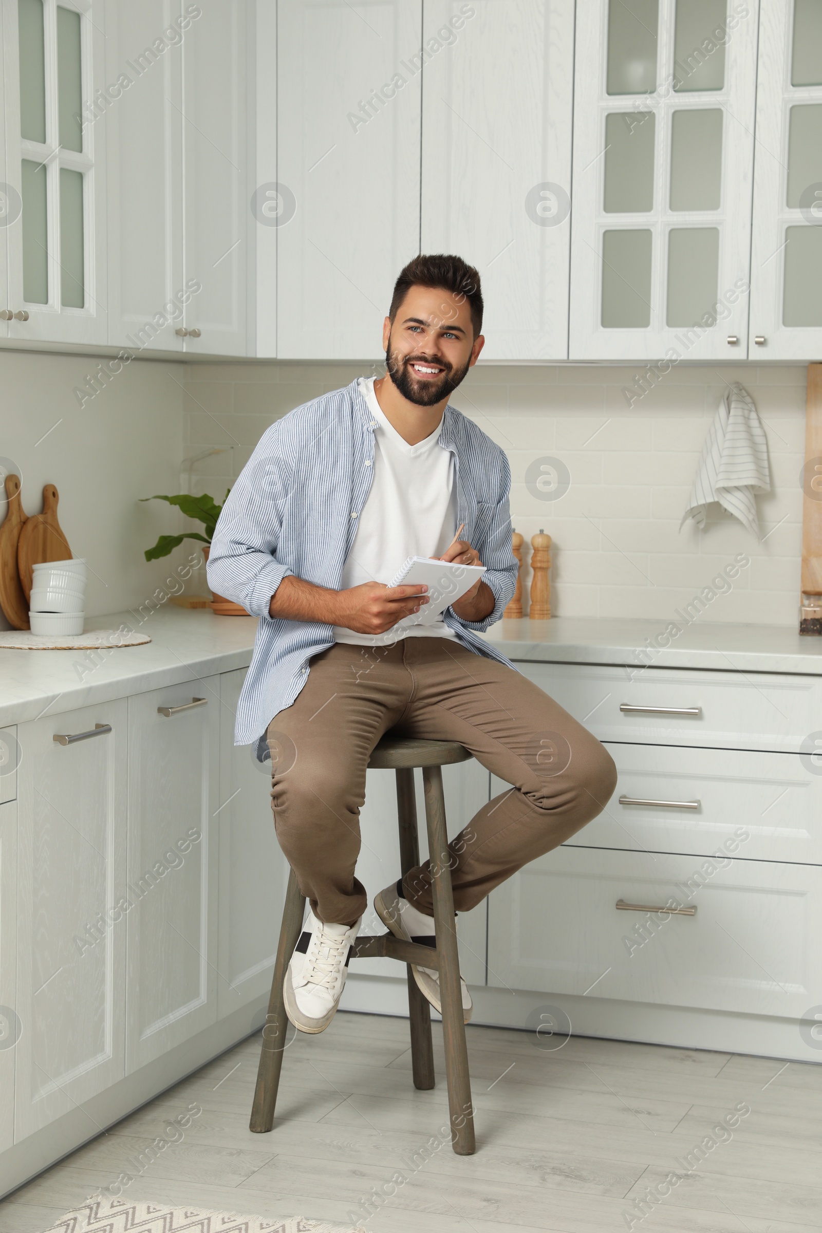 Photo of Handsome young man with notebook sitting on stool in kitchen