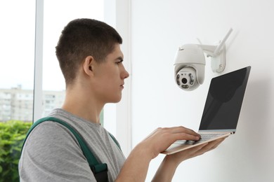 Technician with laptop installing CCTV camera on wall indoors