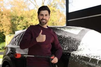 Man with high pressure water jet showing thumbs up near auto at outdoor car wash