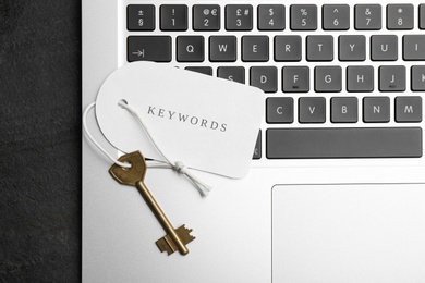 Laptop, key and tag with word KEYWORDS on black table, top view