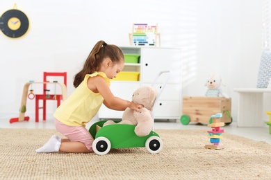 Photo of Cute little girl playing with toy walker and teddy bear on floor at home