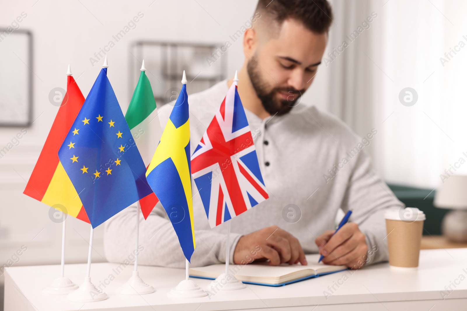 Photo of Young man writing in notebook at white table indoors, focus on different flags