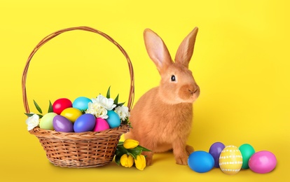 Image of Cute bunny and wicker basket with bright Easter eggs on yellow background