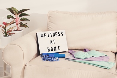 Photo of Sport equipment and lightbox with hashtag FITNESS AT HOME on sofa indoors. Message to promote self-isolation during COVID‑19 pandemic