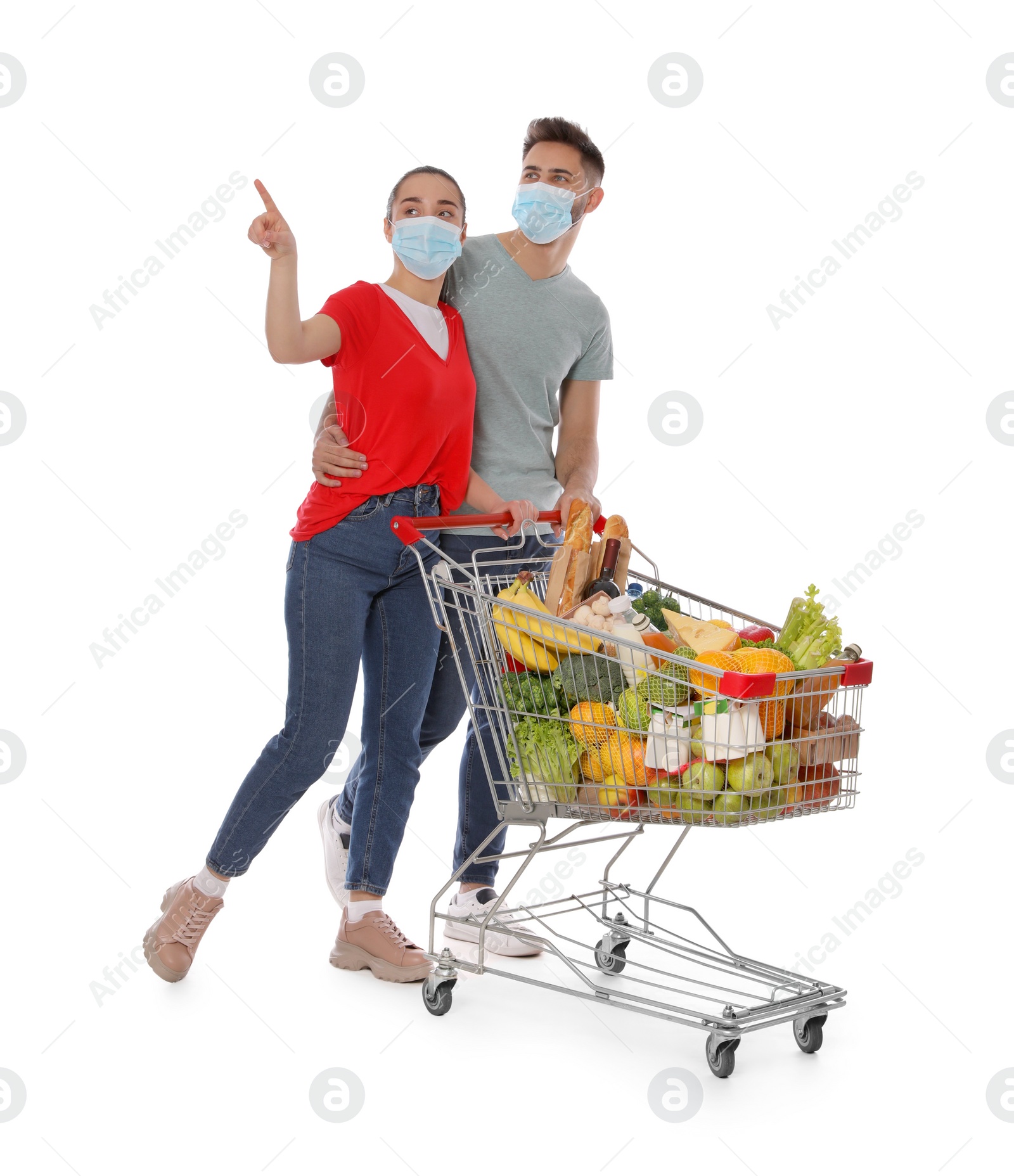 Photo of Couple with protective masks and shopping cart full of groceries on white background