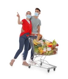 Couple with protective masks and shopping cart full of groceries on white background