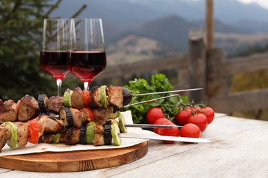 Photo of Metal skewers with delicious meat, vegetables and wine served on wooden table against mountain landscape. Space for text