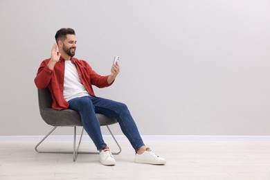 Photo of Handsome man having video chat via smartphone while sitting in armchair near light grey wall indoors, space for text