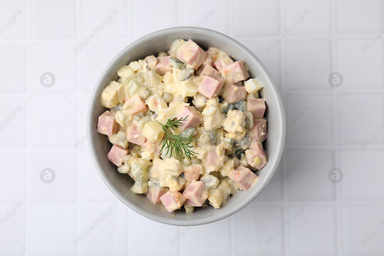 Photo of Tasty Olivier salad with boiled sausage in bowl on white tiled table, top view