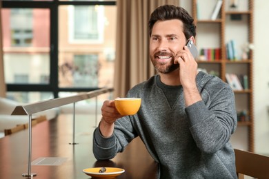 Photo of Handsome man with cup of coffee talking on phone at table in cafe