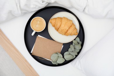 Photo of Tray with tasty croissant, cup of coffee and envelope on white bed, top view
