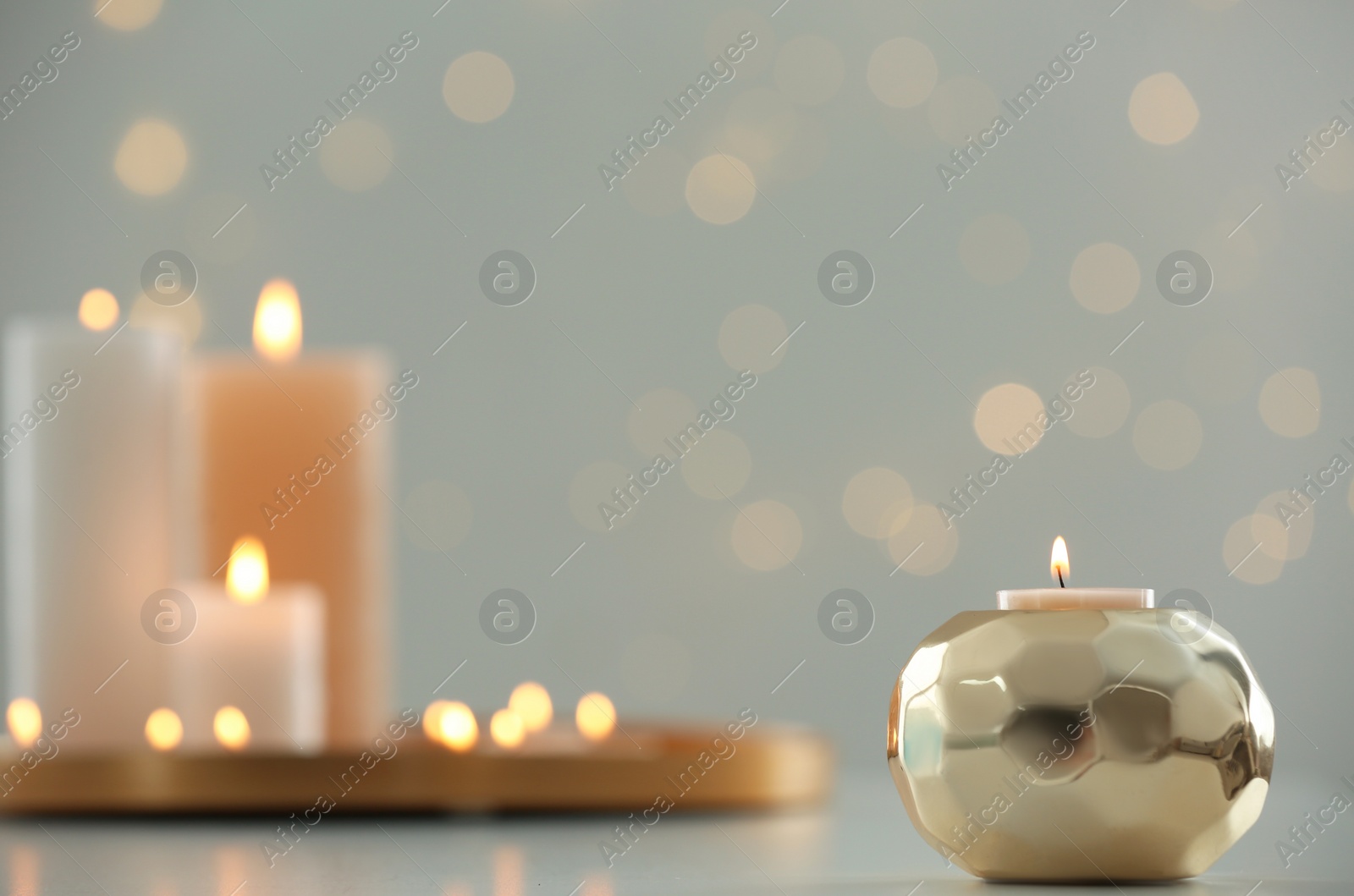 Photo of Burning candle in golden holder on table against blurred background with bokeh effect, closeup. Space for text