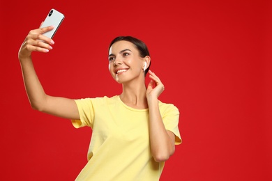 Photo of Happy young woman with wireless earphones taking selfie on red background