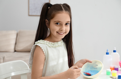 Photo of Cute little girl mixing ingredients with silicone spatula at table in room. DIY slime toy
