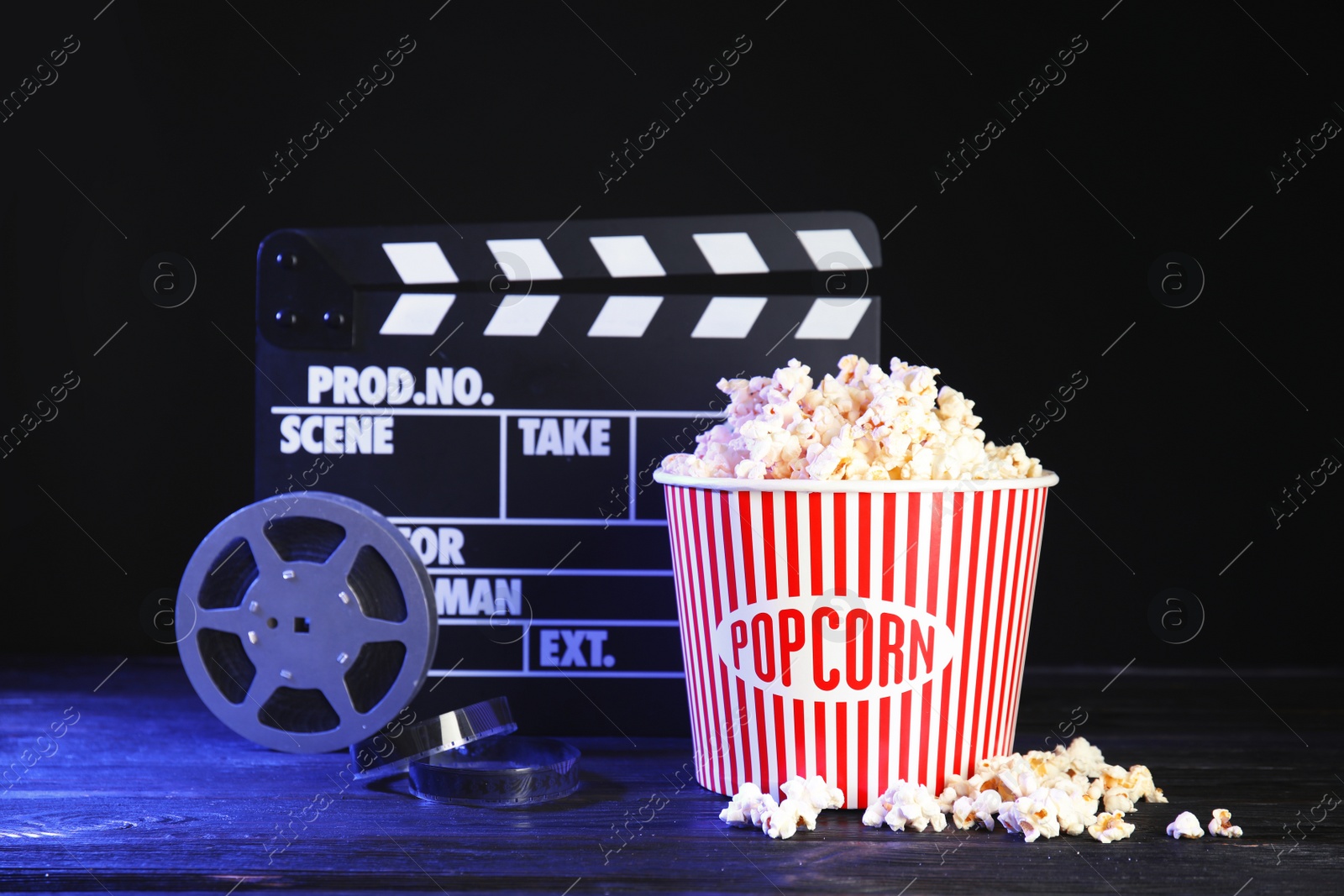 Photo of Clapperboard, reel and popcorn bucket on table against dark background. Watching cinema