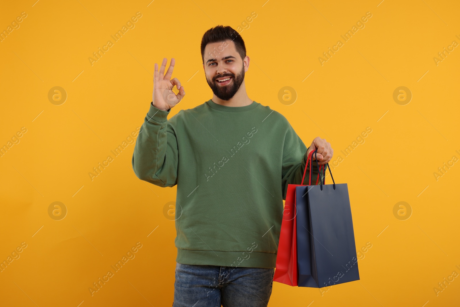 Photo of Happy man with many paper shopping bags showing ok gesture on orange background