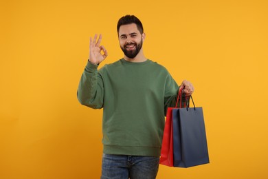 Photo of Happy man with many paper shopping bags showing ok gesture on orange background