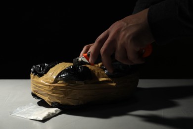 Smuggling and drug trafficking. Man opening package of narcotics with box cutter at light table against black background, closeup