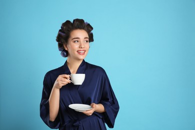 Photo of Happy young woman in silk bathrobe with hair curlers holding cup of drink on light blue background, space for text