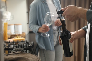 Man opening wine bottle with corkscrew while woman holding wineglass indoors, closeup